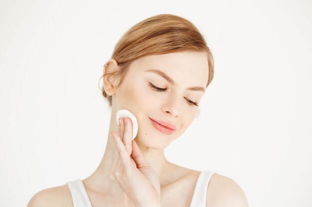 Hermosa chica rubia natural limpieza cara con esponja de algodón sonriendo. Cosmetología salud y spa.