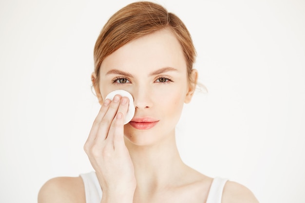 Hermosa chica rubia natural limpieza cara con esponja de algodón sonriendo. Cosmetología y spa.