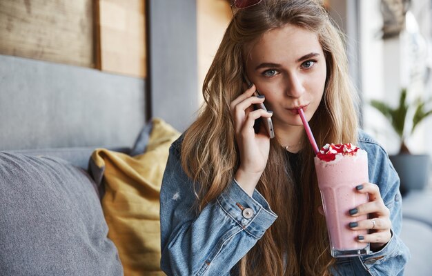 Hermosa chica rubia sentada en la cafetería y bebiendo batido, habla