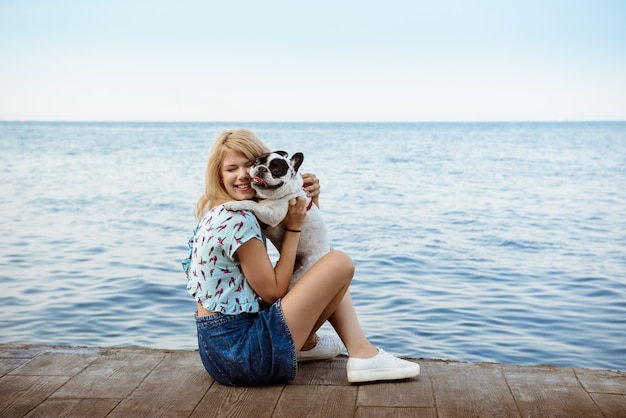 Hermosa chica rubia sentada, jugando con bulldog francés cerca del mar