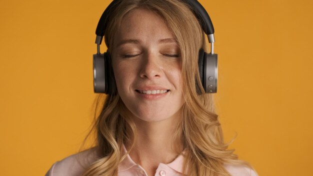 Hermosa chica rubia soñadora escuchando música en auriculares con los ojos cerrados sobre fondo amarillo