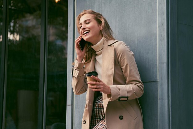 Hermosa chica rubia sonriente en elegante gabardina con café para ir felizmente hablando por teléfono celular al aire libre