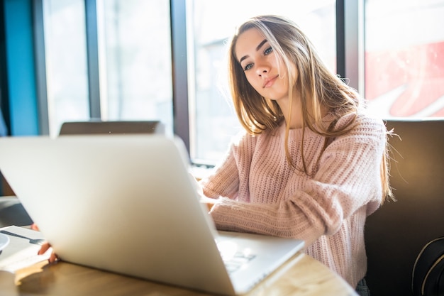 Hermosa chica rubia en suéter blanco de moda trabajando en su computadora portátil en la cafetería en el tiempo diario