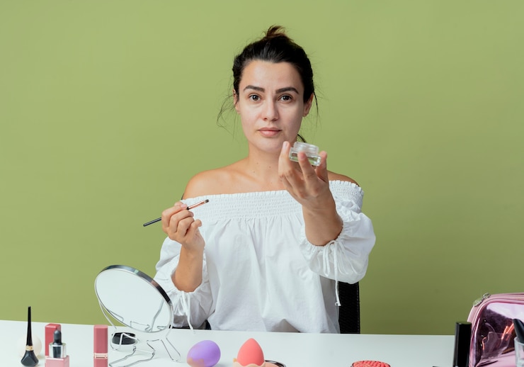 Foto gratuito hermosa chica segura se sienta a la mesa con herramientas de maquillaje con pincel de maquillaje y paleta de sombra de ojos aislada en la pared verde