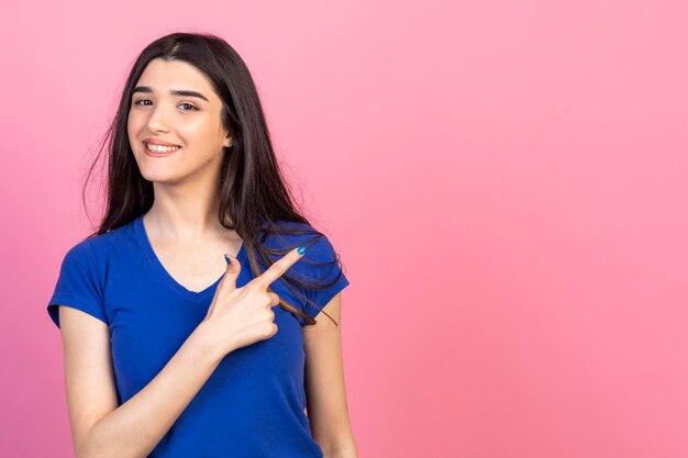 La hermosa chica señala su dedo a un lado y sonríe para la cámara con fondo rosa