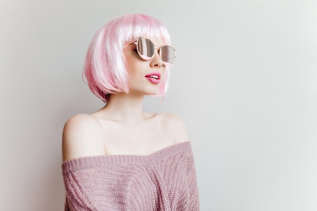 Hermosa chica sensual con peluca rosa posando en la pared de luz. Retrato interior de una mujer bonita en traje púrpura tejido de pie