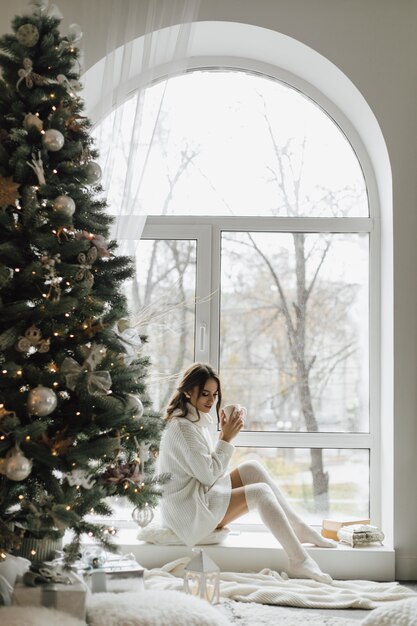 Hermosa chica sentada con una taza de té y cuadros cerca de un árbol de Navidad