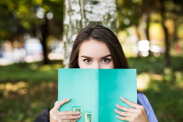 Hermosa chica seria de pelo oscuro en chaqueta de jeans cubre su rostro con un libro contra el parque de verano verde.