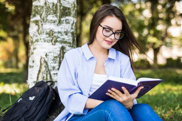 Hermosa chica seria de pelo oscuro en chaqueta de jeans y gafas lee el libro contra el parque verde de verano.