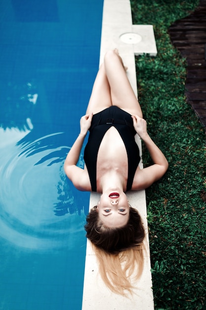 Hermosa chica sexy de lujo se encuentra en el borde de la piscina, tomando el sol en bikini negro relajándose en una isla tropical, una vista superior