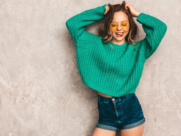 Foto gratuita hermosa chica sexy sonriente hermosa en suéter verde de moda. mujer posando en gafas de sol redondas. modelo divirtiéndose y mostrando su lengua