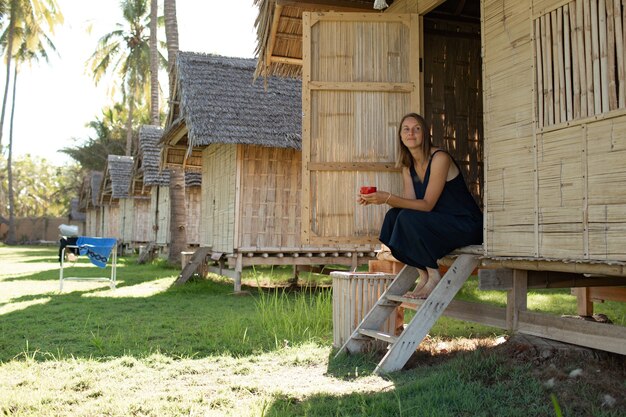 Hermosa chica se sienta cerca del bungalow y bebe café.