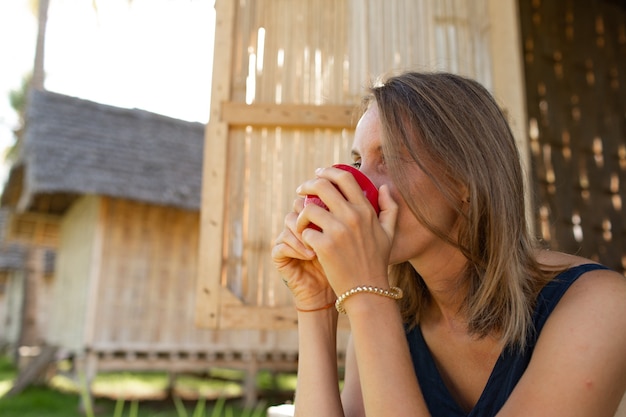 Hermosa chica se sienta cerca del bungalow y bebe café.