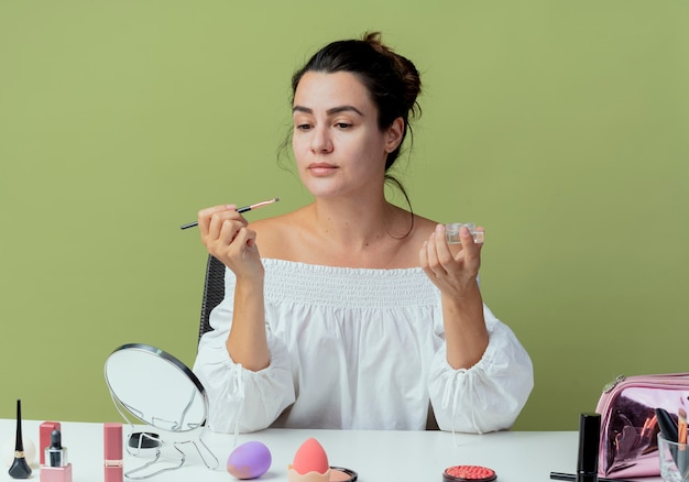 Hermosa chica se sienta a la mesa con herramientas de maquillaje listas para aplicar sombra de ojos con pincel de maquillaje aislado en la pared verde