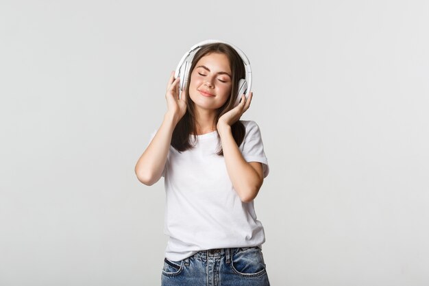 Hermosa chica soñadora disfrutando de escuchar música en auriculares inalámbricos, sonriendo feliz.