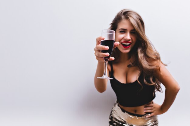 Foto gratuita hermosa chica sonriendo y levantando una copa de vino en un brindis. lleva falda con lentejuelas, top negro. look sexy y elegante con vientre desnudo y escote profundo. centrarse en una copa de vino tinto. aislado.