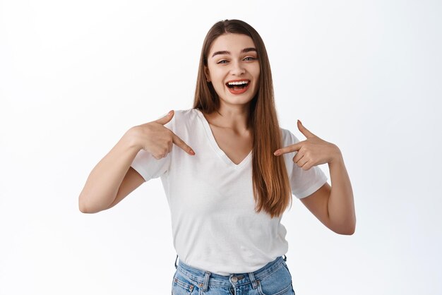 Hermosa chica sonriente apuntándose a sí misma con cara segura de sí misma autopromocionando su voluntariado de pie sobre fondo blanco Copiar espacio