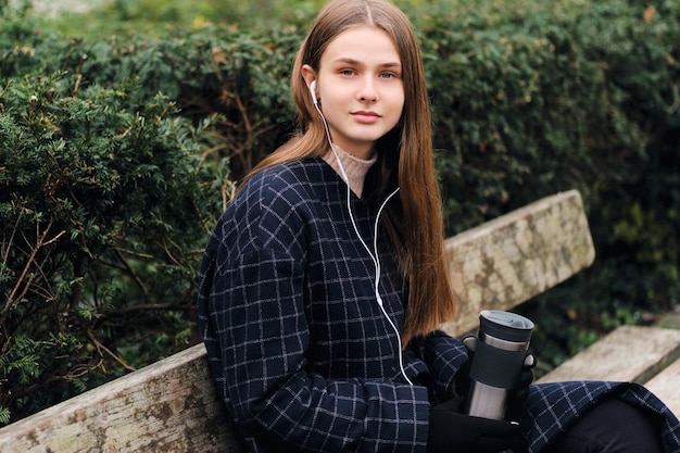 Hermosa chica sonriente en auriculares sosteniendo una taza termo mirando con confianza a la cámara en un banco en el parque de la ciudad
