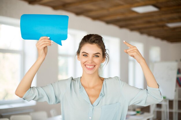 Hermosa chica sonriente en blusa mirando felizmente en la cámara sosteniendo un icono de mensaje de papel azul en la mano cerca de la cabeza mientras pasa tiempo en la oficina