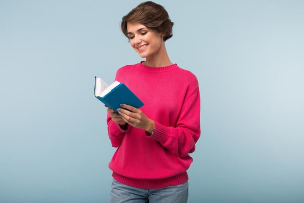 Hermosa chica sonriente con cabello corto oscuro en suéter rosa y jeans leyendo felizmente un pequeño libro sobre fondo azul aislado