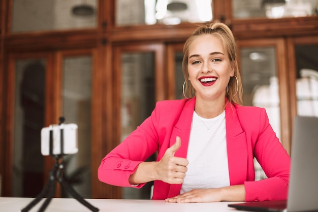 Foto gratuita hermosa chica sonriente con chaqueta rosa mirando felizmente a la cámara mostrando el pulgar hacia arriba mientras graba un nuevo video para vlo