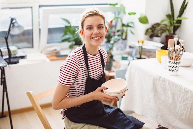 Hermosa chica sonriente con delantal negro y camiseta a rayas sosteniendo cuencos de arcilla hechos a mano en las manos mirando alegremente a la cámara en el moderno estudio de cerámica