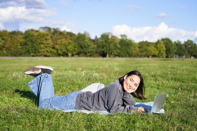 Hermosa chica sonriente descansando en el parque tirada en el césped con una laptop estudiando o viendo videos en línea