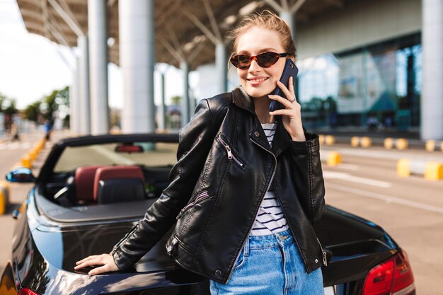 Hermosa chica sonriente con gafas de sol y chaqueta de cuero apoyada en un auto descapotable negro mientras habla alegremente por teléfono celular con el aeropuerto en el fondo