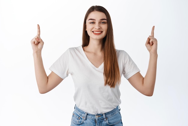 Hermosa chica sonriente que muestra con orgullo un anuncio que señala con el dedo hacia arriba en la mejor oferta promocional muestra descuentos en la tienda de pie sobre fondo blanco en camiseta