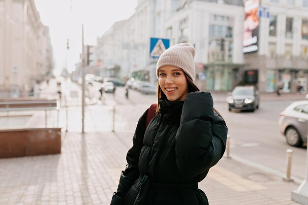 Hermosa chica sonriente con sombrero y chaqueta está mirando a la cámara en el centro de la ciudad a la luz del sol