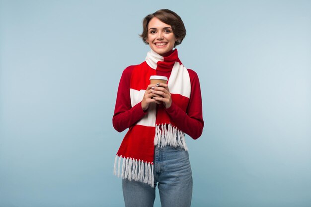 Hermosa chica sonriente en suéter rojo y bufanda sosteniendo una taza de café para ir de la mano mientras mira felizmente en cámara sobre fondo azul.