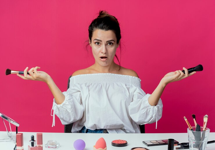 Foto gratuito hermosa chica sorprendida se sienta a la mesa con herramientas de maquillaje sosteniendo pinceles de maquillaje aislados en la pared rosa