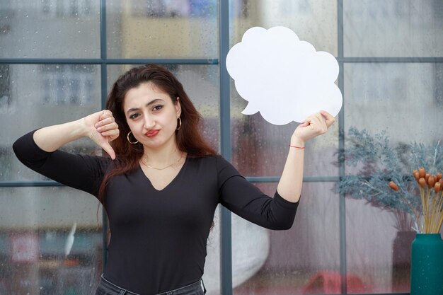 Hermosa chica sosteniendo una burbuja de ideas en forma de nube y gesticulando con el pulgar hacia abajo Foto de alta calidad