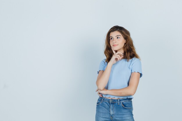 Hermosa chica sosteniendo el dedo en la barbilla en camiseta, jeans y mirando confiado. vista frontal.