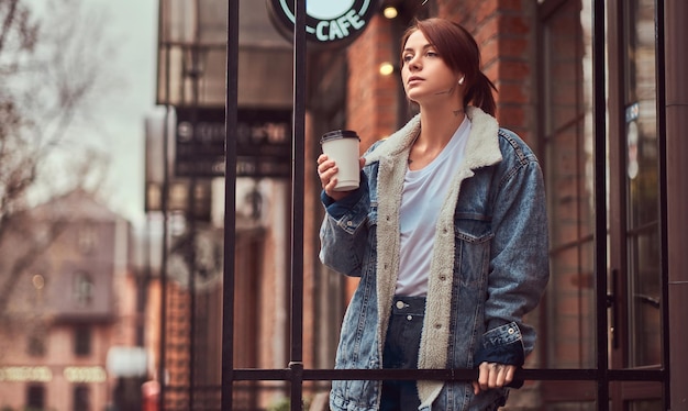 Una hermosa chica tatuada con un abrigo de mezclilla sosteniendo una taza con café para llevar afuera cerca del café.