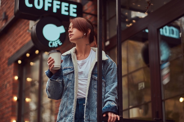 Una hermosa chica tatuada con un abrigo de mezclilla sosteniendo una taza con café para llevar afuera cerca del café.