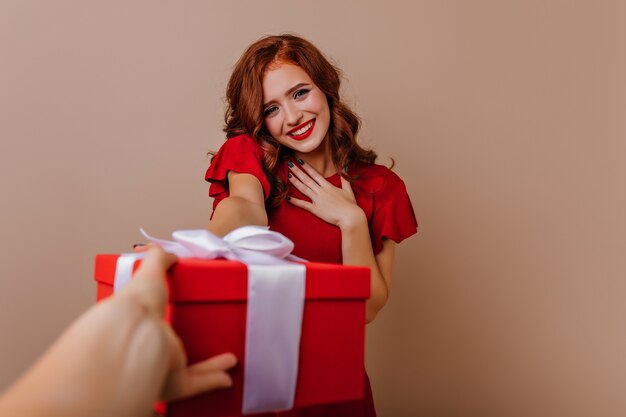 Hermosa chica tímida en vestido rojo posando en su cumpleaños. Foto interior de mujer de inspiración rizada con regalo de año nuevo.