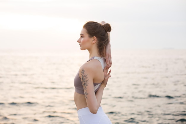 Hermosa chica en top deportivo y polainas blancas cogidos de la mano detrás de la espalda con vista al mar en el fondo. Mujer joven mirando soñadoramente a un lado mientras practica yoga junto al mar