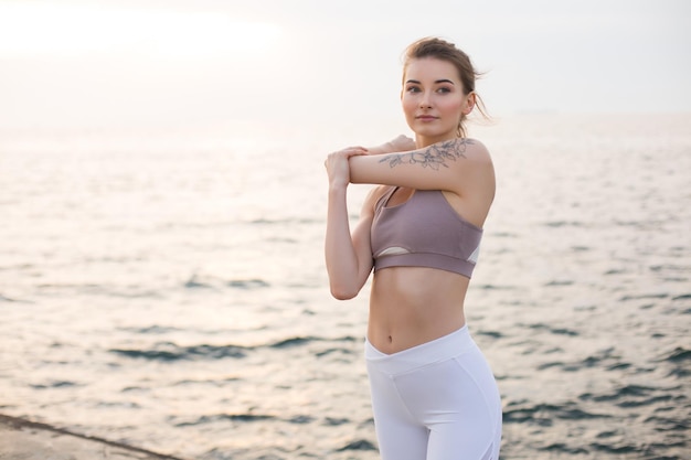 Hermosa chica en top deportivo y polainas blancas practicando yoga con hermosa vista al mar en el fondo. Mujer joven mirando cuidadosamente a un lado mientras se estira junto al mar