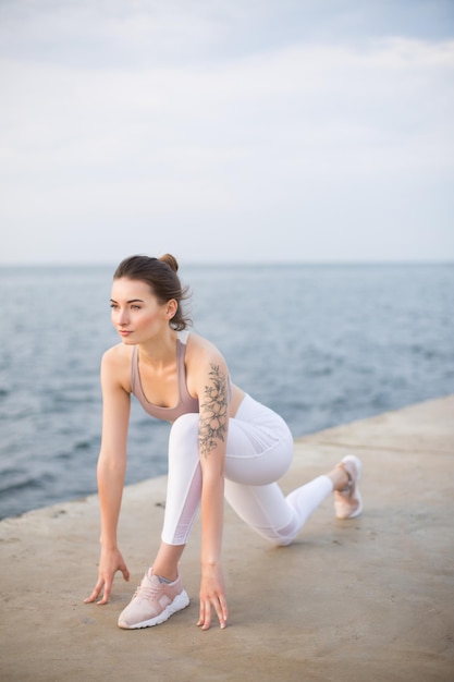 Hermosa chica en top deportivo y polainas blancas practicando yoga con vista al mar en el fondo. Mujer joven mirando cuidadosamente a un lado mientras entrena junto al mar