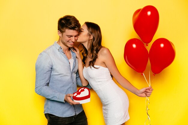 Hermosa chica vestida de blanco con globos rojos besando a su novio