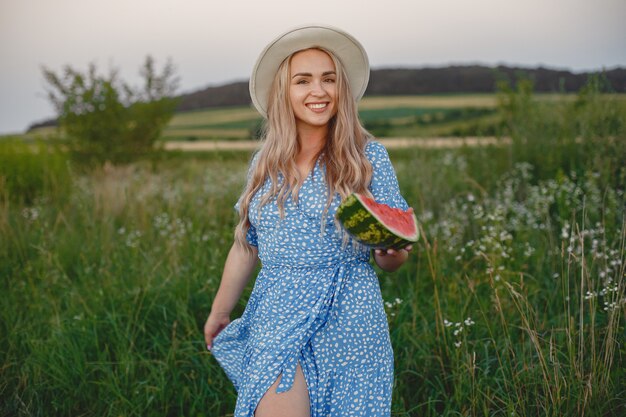 Hermosa chica con un vestido azul y un sombrero. Mujer en un campo de verano. Chica con una sandía.