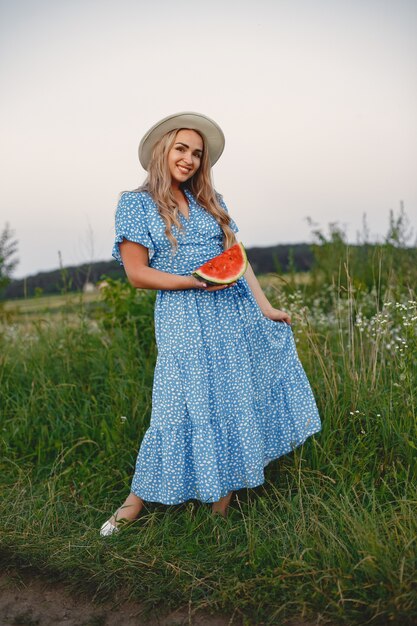 Hermosa chica con un vestido azul y un sombrero. Mujer en un campo de verano. Chica con una sandía.