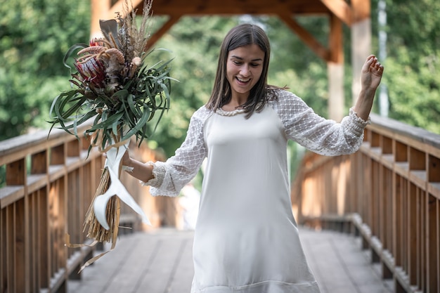 Hermosa chica con un vestido blanco con un ramo de flores exóticas en un puente de madera.