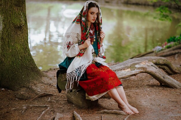 Hermosa chica en un vestido étnico tradicional sentado en un banco cerca del lago