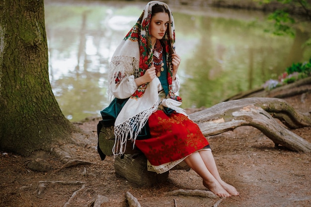 Hermosa chica en un vestido étnico tradicional sentado en un banco cerca del lago