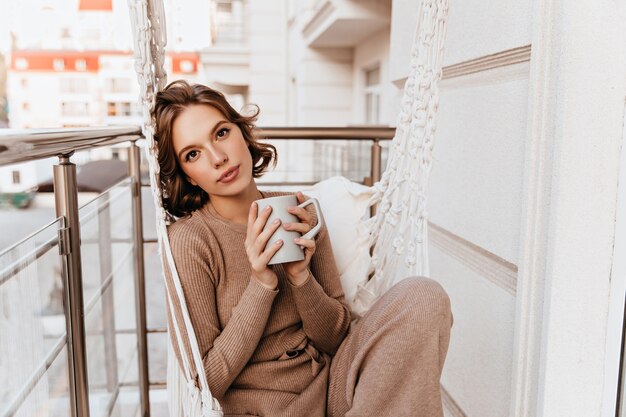 Hermosa chica en vestido de punto tomando café en la mañana. Mujer joven caucásica romántica que sostiene la taza de té en el balcón.