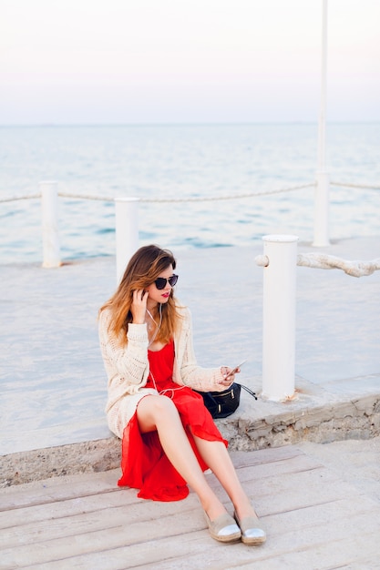 Hermosa chica en vestido rojo y chaqueta blanca se sienta en un muelle, sonríe y escucha música con auriculares en un teléfono inteligente.