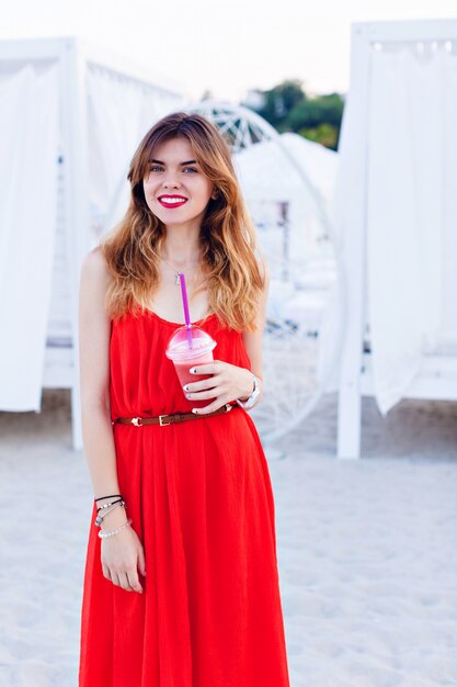 Hermosa chica en vestido rojo de pie en una playa y sonriendo ampliamente