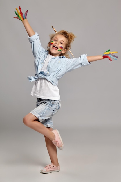 Hermosa colegiala con un cepillo en su elegante cabello rubio rizado, vestida con una camisa azul y una camiseta blanca. Ella está posando con manos y mejillas pintadas, sonriendo y luciendo muy feliz, sobre un fondo gris.